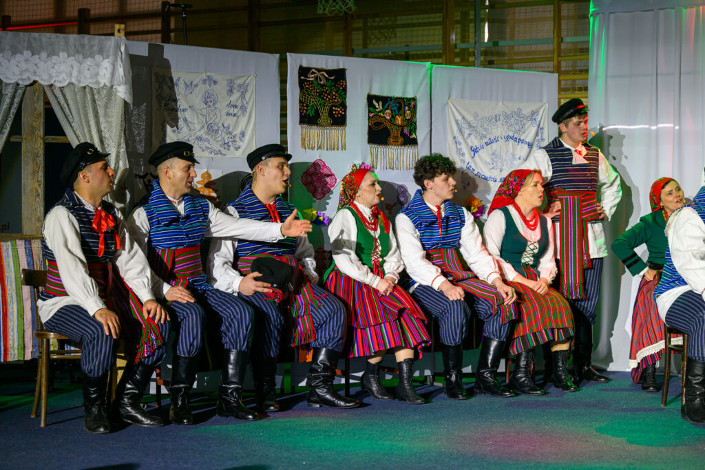 Grupa ludzi w strojach ludowych siedzi na scenie. Kolorowe wzory i hafty zdobią tło. Na twarzach widać skupienie.