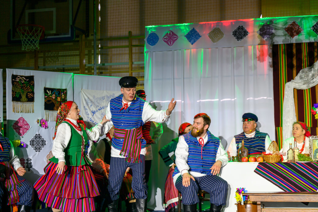 Grupa osób w stroju ludowym na scenie, ubrana w kolorowe ubrania. Tło zdobią białe zasłony i haftowane dekoracje. Na stole znajdują się kosze z owocami i butelki.