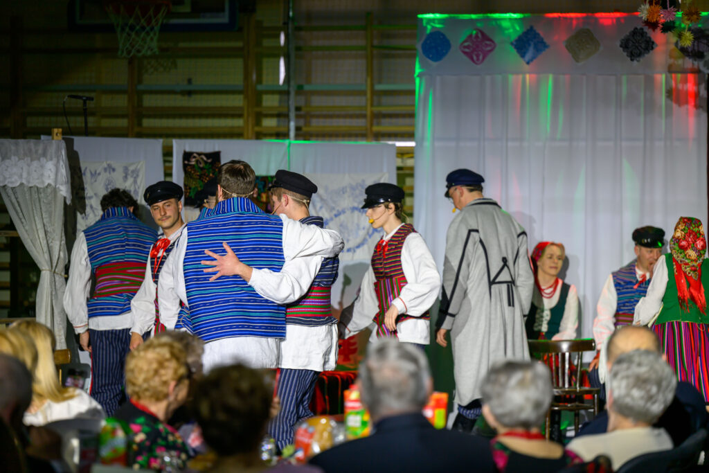 Grupa osób w kolorowych strojach ludowych tańczy na scenie. Tło zdobią białe zasłony i dekoracje. Przed sceną siedzą widzowie.