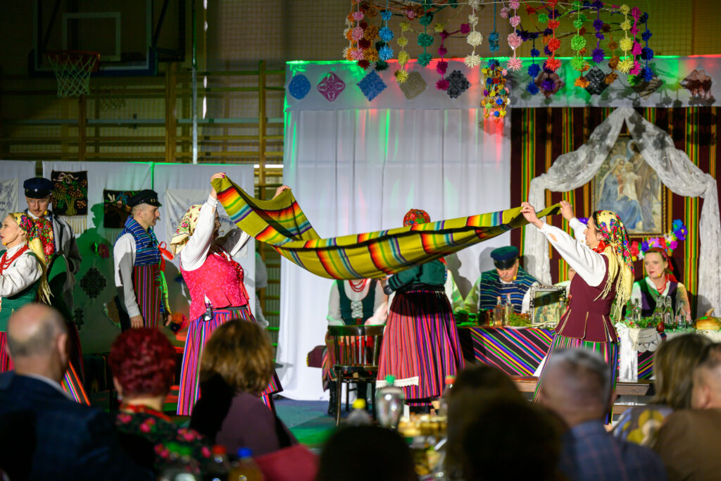 Osoby w strojach ludowych trzymają kolorowy szal na tle dekoracji i postaci w sali. Ściany zdobią białe i kolorowe tkaniny, scena jest żywa. Na pierwszym planie widoczna jest publiczność w rozmazanych kolorach.