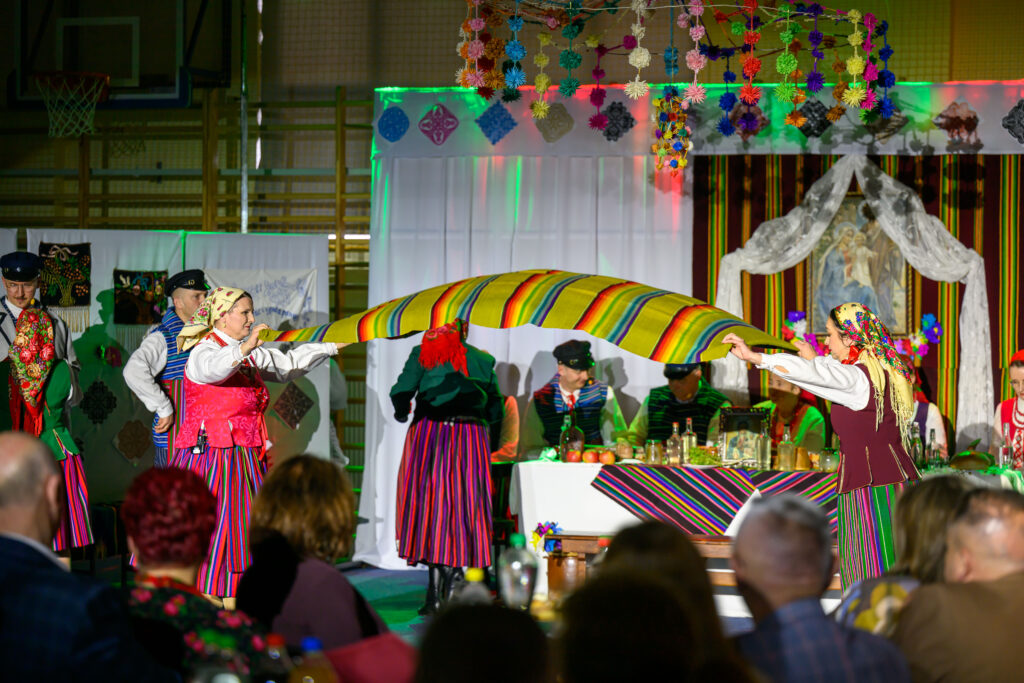 Ludzie w tradycyjnych strojach ludowych tańczą z kolorową tkaniną. Tło zdobione girlandami i obrazem świętym. Oświetlenie sceniczne w ciepłych barwach.