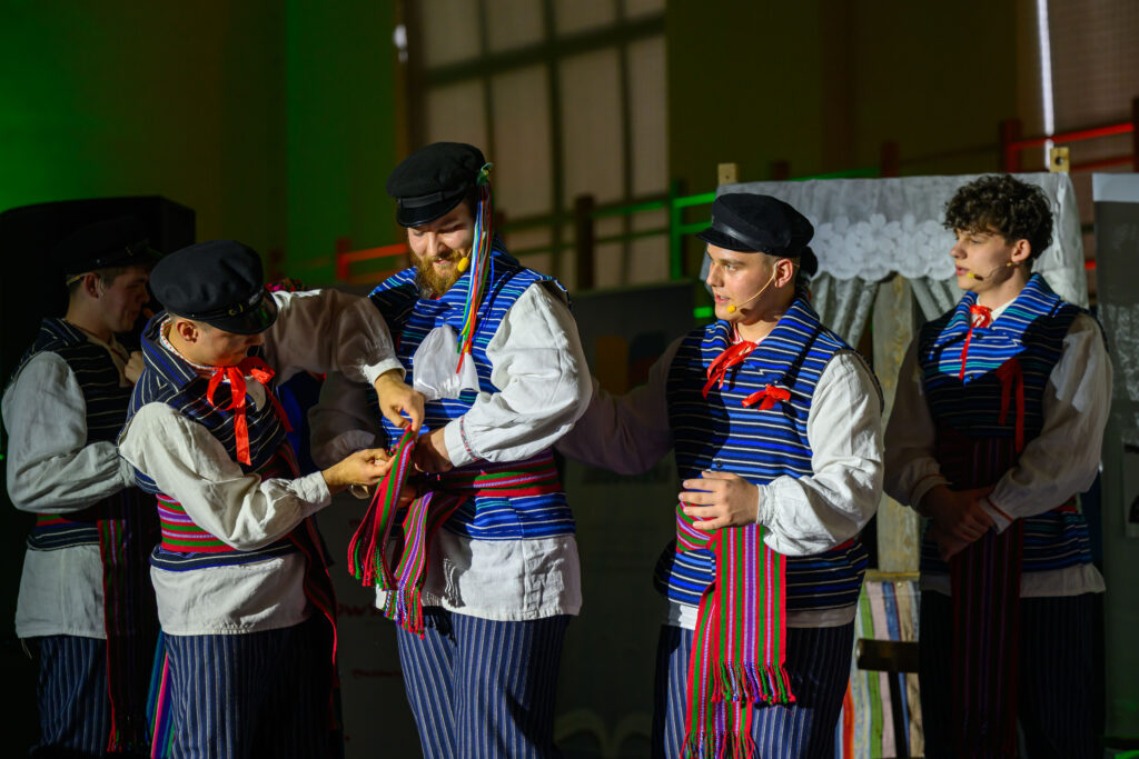 Grupa mężczyzn w strojach ludowych, z elementami niebieskimi i czerwonymi. Tło jest ciemne z dekoracjami. Intensywne oświetlenie na scenie.