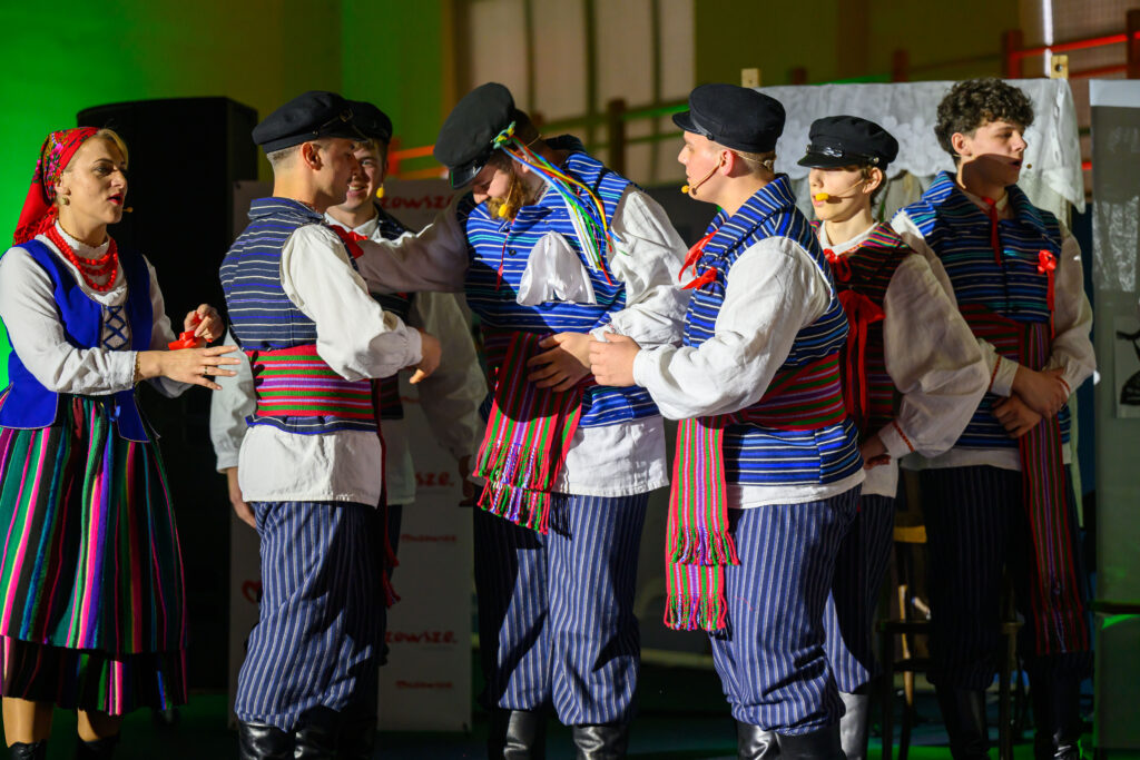 Grupa osób w tradycyjnych strojach ludowych. Kolorowe pasy i kapelusze. Tło oświetlone zielonym światłem.