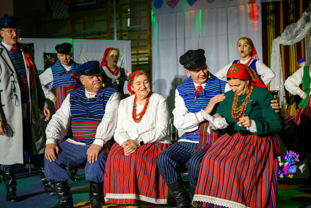 Grupa ludzi w tradycyjnych strojach ludowych siedzi na scenie. Kolorowe ubrania z dominacją czerwieni i niebieskiego. Tło to jasne płótno z dekoracjami.