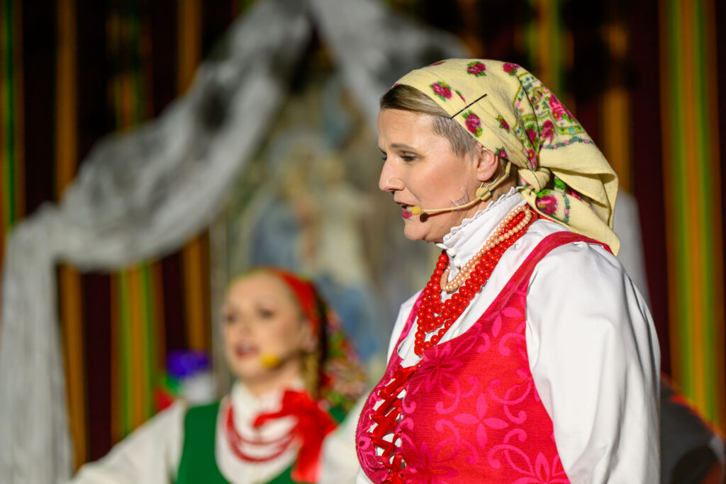 Kobieta w kolorowym stroju ludowym z chustą i koralami występuje na scenie. Tło jest ozdobione wzorzystymi tkaninami. Druga osoba w tle w podobnym stroju.