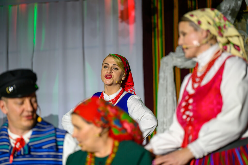 Grupa osób w kolorowych strojach ludowych na scenie. Przeważają kolory czerwony, niebieski i zielony. Za nimi widać białe zasłony.