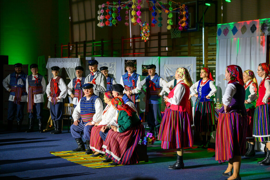 Grupa ludzi w kolorowych strojach ludowych tańczy na scenie. Tło jest zielone z dekoracyjnym, wiszącym ornamentem. Scena jest jasna i ozdobiona kolorami.