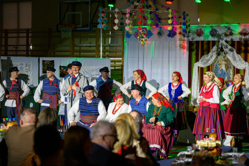 Grupa ludzi w strojach ludowych na scenie, kolorowe dekoracje wiszące z sufitu, tło z jasnym oświetleniem.