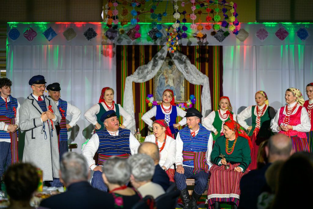 Grupa ludzi w strojach ludowych na scenie; kolorowe pasy, czerwone i niebieskie elementy. Tło ozdobione papierowymi dekoracjami. W centrum obraz z welonem.
