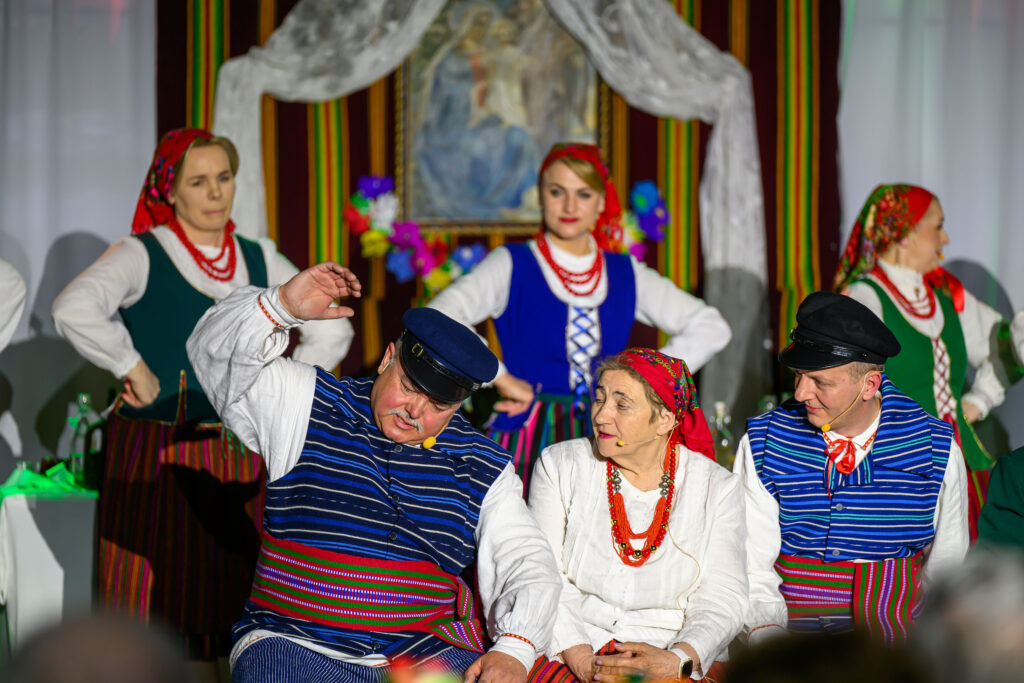 Na zdjęciu widać ludzi w tradycyjnych ludowych strojach. Kolorowe pasy dominują w tkaninach, a w tle znajduje się ozdobny obraz. Grupa jest na scenie, co sugeruje występ.