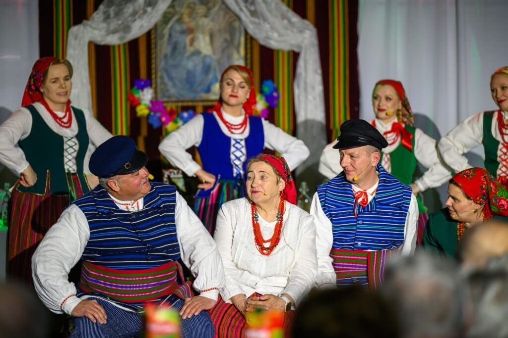 Grupa osób w tradycyjnych strojach ludowych siedzi i stoi na scenie. Kolory strojów to niebieski, czerwony i biały. Tło zdobią pasiaste zasłony i obraz.