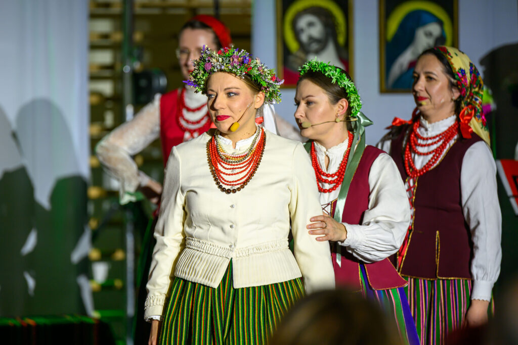 Na zdjęciu cztery kobiety w tradycyjnych strojach ludowych. Wyróżniają się kwieciste wianki i kolorowe korale. Tło stanowią obrazy religijne.
