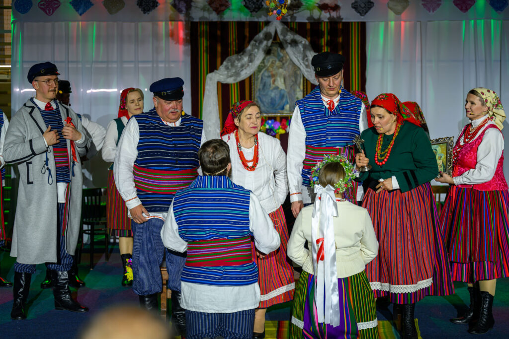 Grupa osób w kolorowych, ludowych strojach stoi razem na tle jasnej sceny. Dominują odcienie niebieskiego, czerwonego i białego. W centrum jest kobieta z kwiecistym wiankiem.