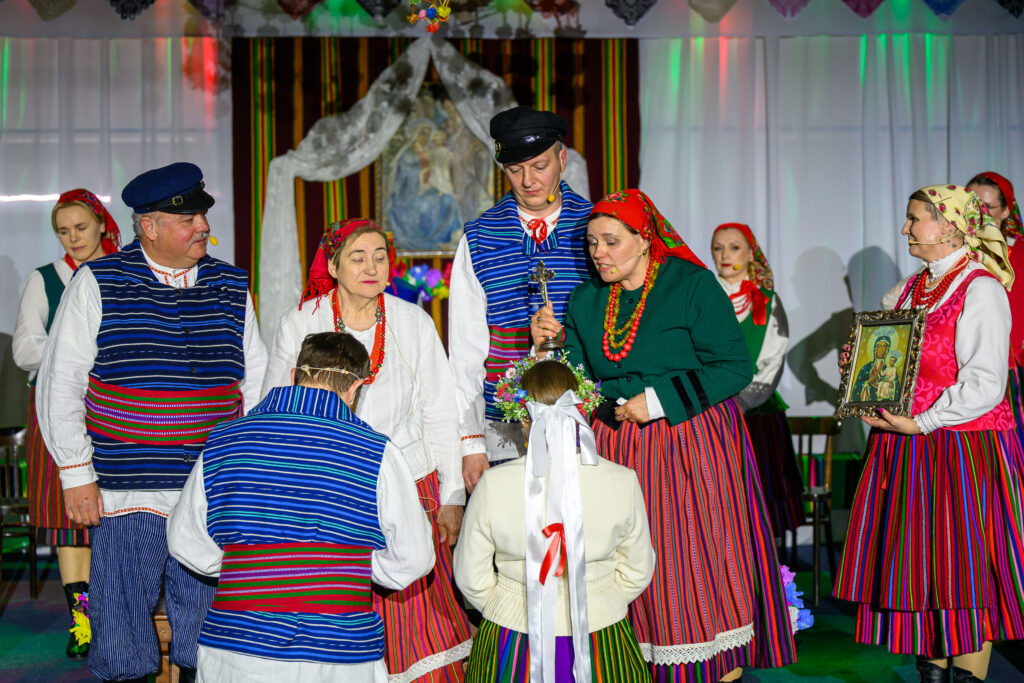 Grupa osób w tradycyjnych, kolorowych strojach ludowych stoi na scenie. Tło jest białe z kolorowymi światłami. Kobieta trzyma obrazek religijny.