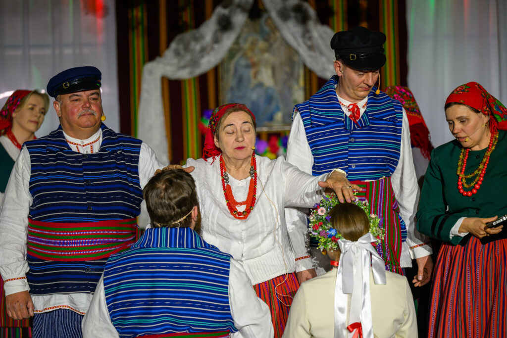 Ludzie w kolorowych strojach ludowych uczestniczą w uroczystości. Barwy dominujące to niebieski, czerwony i biały. Tło jest ozdobione tradycyjnymi wzorami i materiałami.