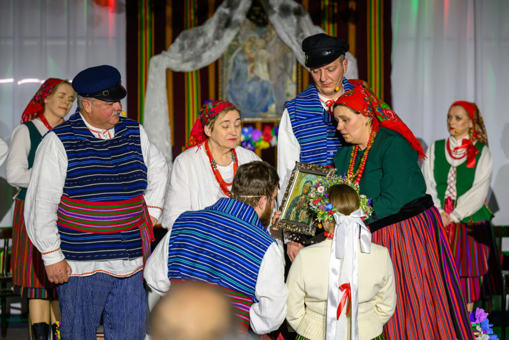 Ubrani w kolorowe stroje ludowe ludzie trzymają obraz. Tło zdobią pasiaste zasłony i biały materiał. Na podłodze leżą kwiaty.