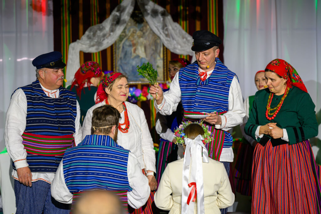 Grupa osób w barwnych strojach ludowych na scenie. Dominuje kolor niebieski, biały i czerwony. W tle tkaniny i obraz.