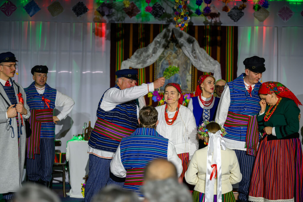 Grupa ludzi w tradycyjnych strojach ludowych na scenie. Kobiety w kolorowych spódnicach i chustach, mężczyźni w pasiakach. Tło ozdobione kolorowymi dekoracjami.