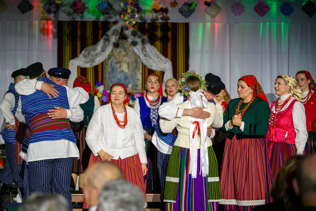 Na zdjęciu grupa osób w kolorowych, tradycyjnych strojach ludowych. Tło zdobią barwne dekoracje i białe zasłony. Dwójka ludzi przytula się pośrodku sceny.