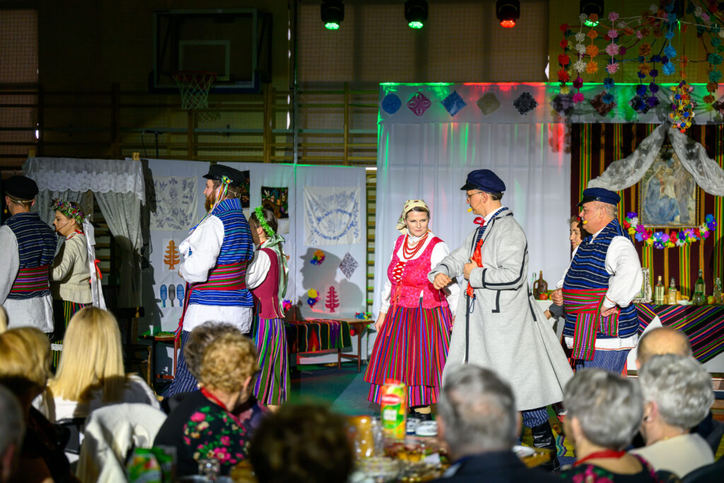 Osoby w strojach ludowych tańczą w kolorowej sali. Stroje mają czerwone, niebieskie i zielone elementy. Tło zdobią dekoracje i wzory.