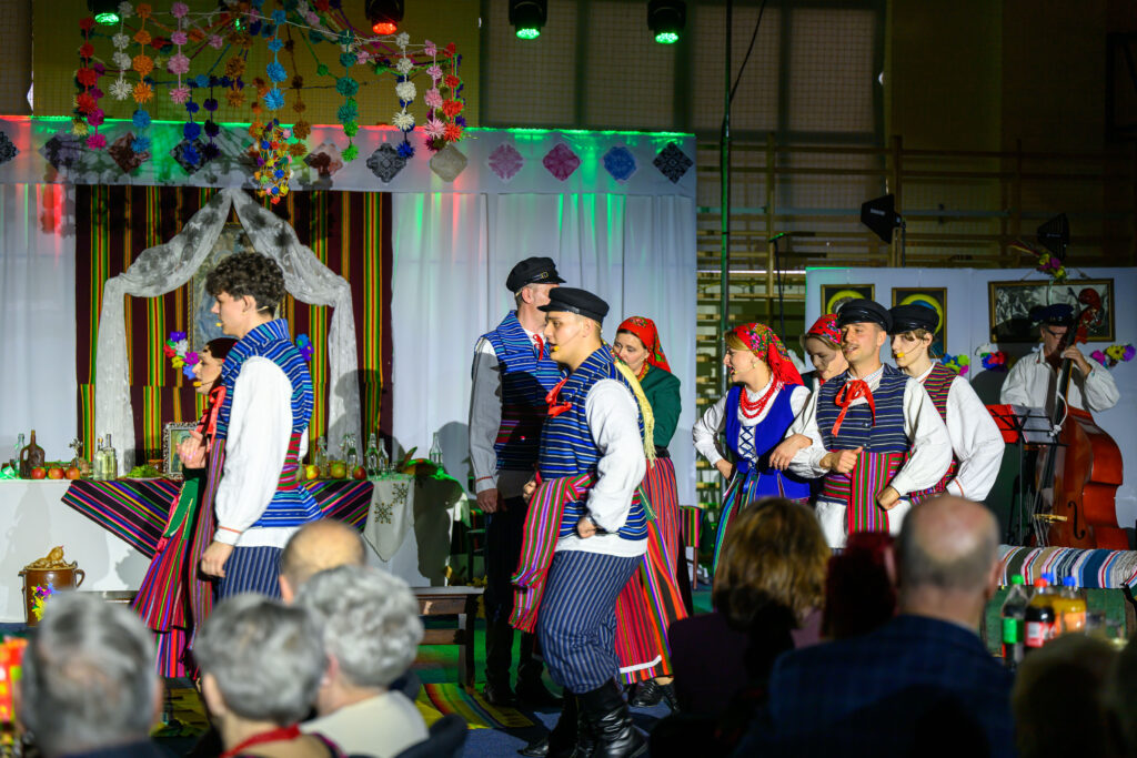 Grupa ludzi w kolorowych strojach ludowych tańczy na scenie. Tło jest ozdobione białymi i kolorowymi dekoracjami. Scena oświetlona jest różnobarwnymi światłami.