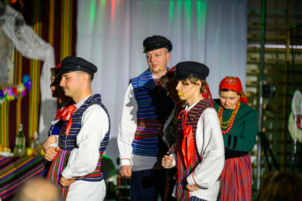 Grupa osób w tradycyjnych strojach ludowych, we wzorzystych kamizelkach i czerwonych chustach, występuje na tle sceny. Tło jest kolorowe, z pionowymi pasami w ciepłych barwach. Światło sceniczne dodaje intensywności kolorom ich