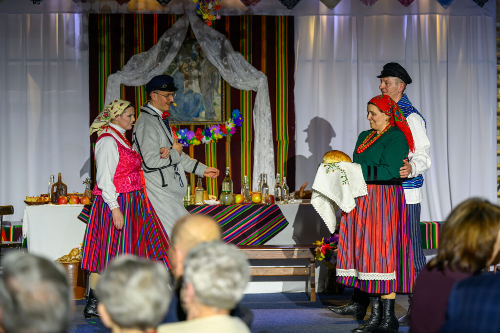 Cztery osoby w stroju ludowym na scenie, trzymają chleb. Kolorowe tło z pionowymi paskami i białymi zasłonami. Na stole stoją napoje i owoce.