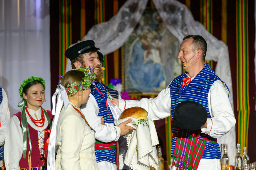 Grupa ludzi w strojach ludowych, z wieńcami i chlebem na białym obrusie. Tło jest kolorowe z obrazem i zasłonami. Dominują barwy niebiesko-czerwone.