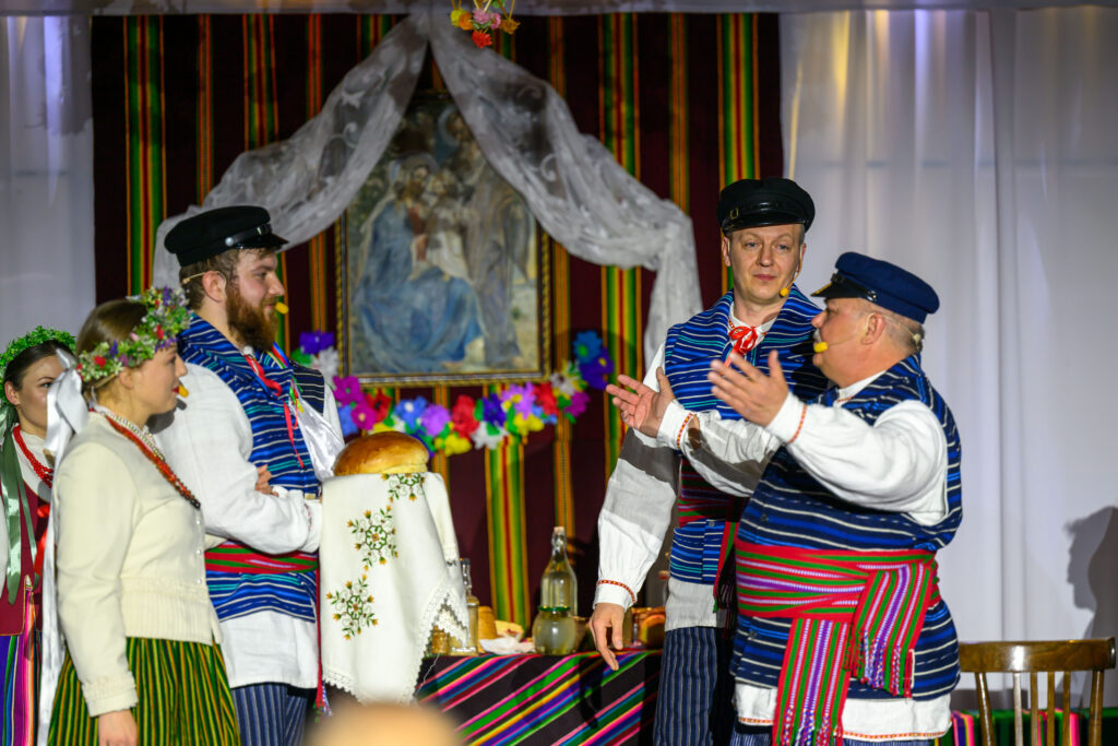 Grupa ludzi w strojach ludowych bierze udział w tradycyjnym obrzędzie. Ubrani są w pasiaste stroje, korony z kwiatów i czapki. W tle widać kolorowe dekoracje i obraz na ścianie.