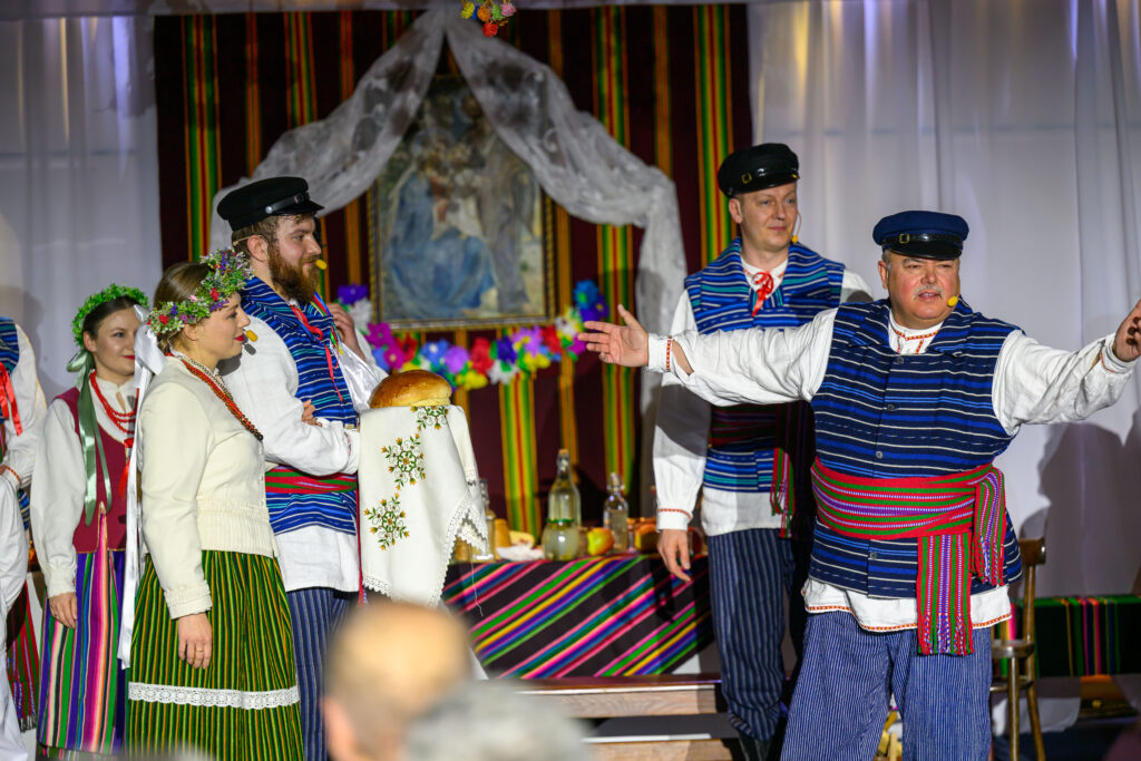 Na zdjęciu są osoby w strojach ludowych z wieńcami i chlebem na białym obrusie. Tło to kolorowe pasy i obraz. Atmosfera jest świąteczna.