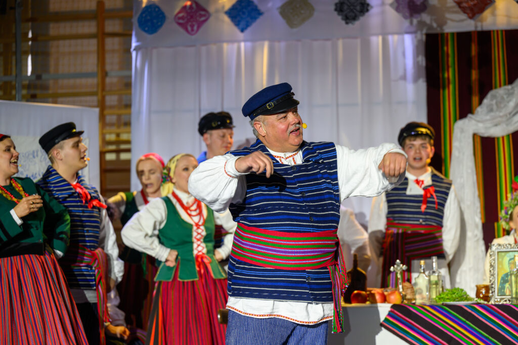 Grupa ludzi w strojach ludowych tańczy na scenie, dominują kolory niebieski, czerwony i zielony. Tło zawiera białe zasłony i kolorowe ozdoby. Na stole obok widoczne są owoce i dekoracje.