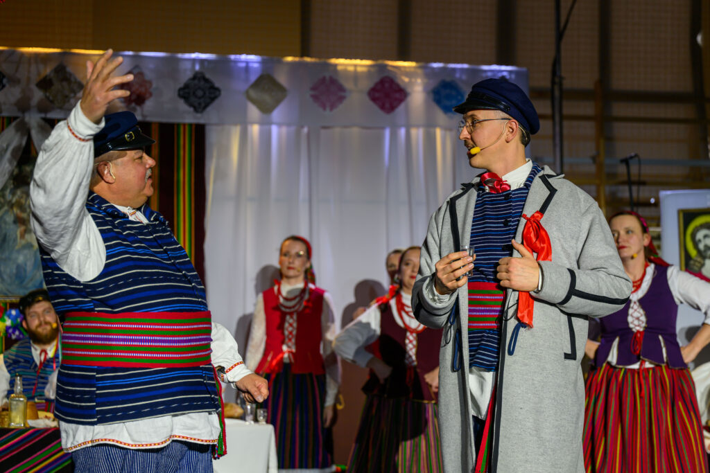 Ludzie w kolorowych strojach ludowych na scenie, na jasnym tle. Dwóch mężczyzn z kapeluszami gestykuluje. W tle kobiety w czerwonych kamizelkach.