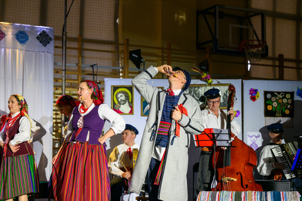 Grupa ludzi w tradycyjnych strojach ludowych występuje na scenie. Ubrania są kolorowe, dominują różnorodne paski i czerwienie. W tle są obrazy religijne i instrumenty muzyczne.