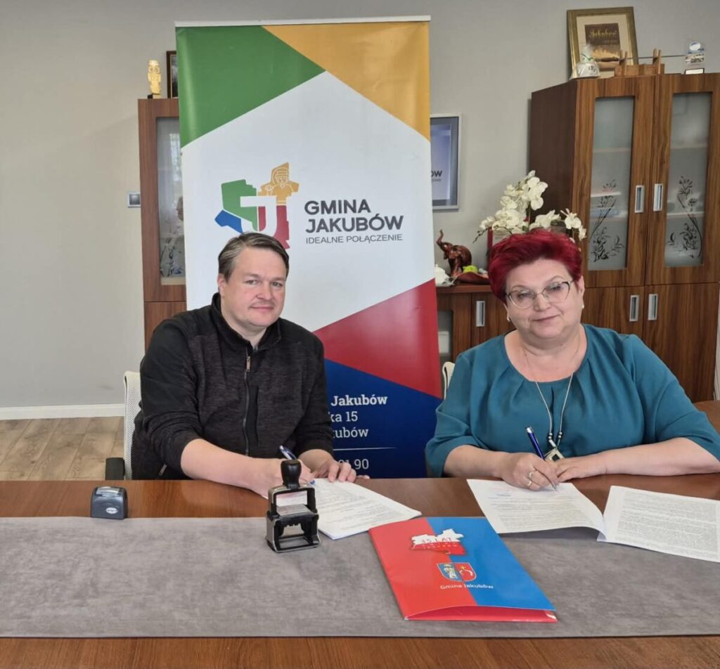 Na zdjęciu siedzi mężczyzna i kobieta przy stole, podpisując dokumenty. Za nimi stoi kolorowy baner z logo gminy. Tło to jasne ściany i drewniana szafka.