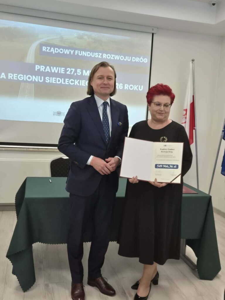 Osoby stoją na tle napisu o funduszu drogowym. Mężczyzna w garniturze i kobieta w czerni trzymają dokument. Na stole zielony obrus.