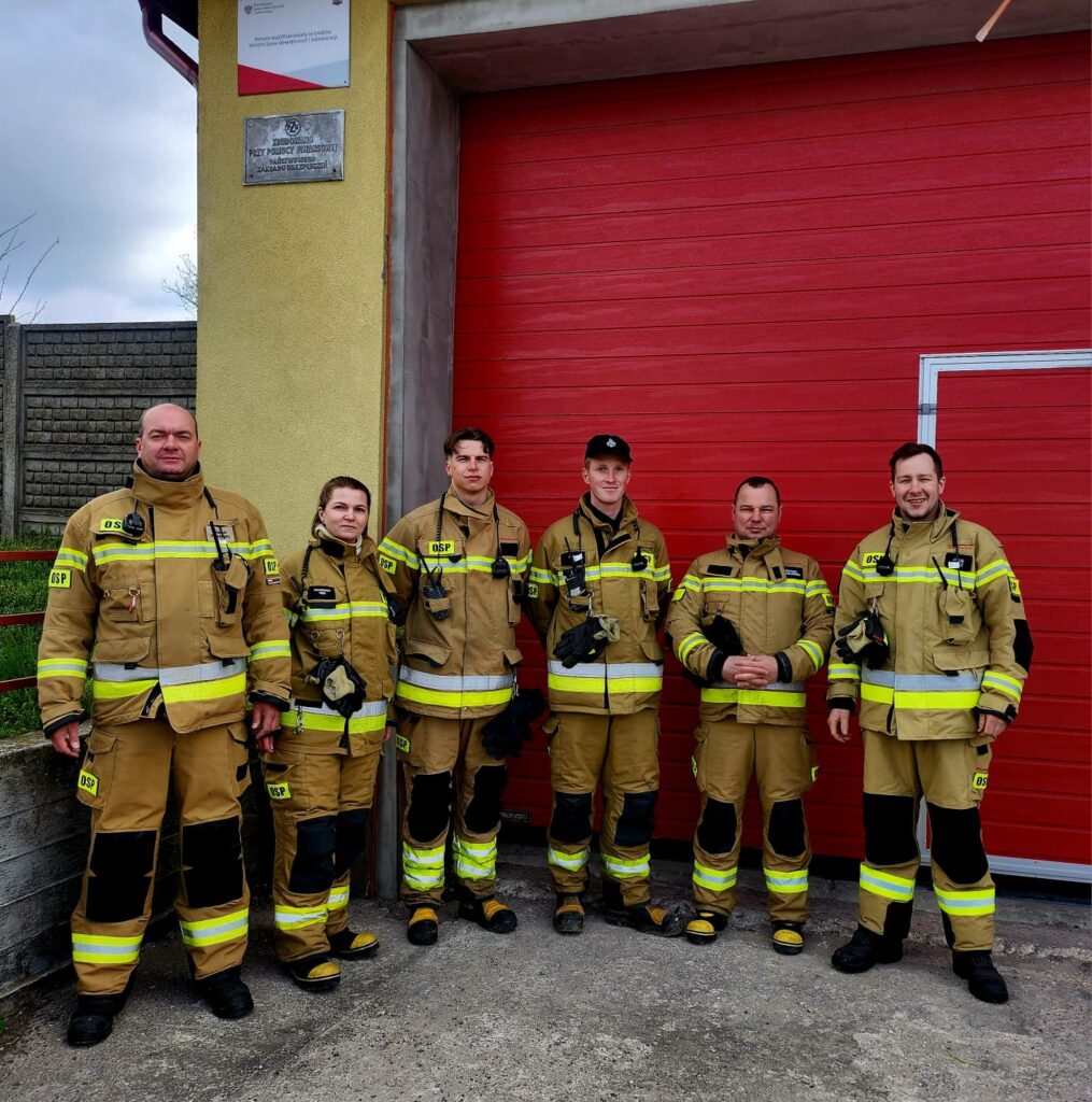 Grupa strażaków w żółto-czarnych mundurach stoi przed czerwoną bramą. Tło jest szare z brązowym murem. Uśmiechają się do kamery.