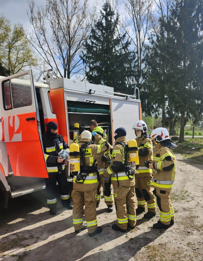 Grupa strażaków w żółtych mundurach stoi przy czerwonym wozie. Na tle widać drzewa i błękitne niebo. Strażacy mają hełmy i sprzęt ochronny.