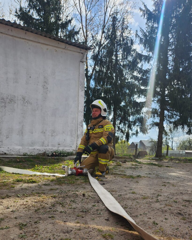 Na zdjęciu strażak w jasnym uniformie klęczy obok białej ściany budynku, obok leży wąż strażacki. Za nim w tle drzewa i niebo z promieniem słońca. Tło jest słoneczne, pełne zieleni.