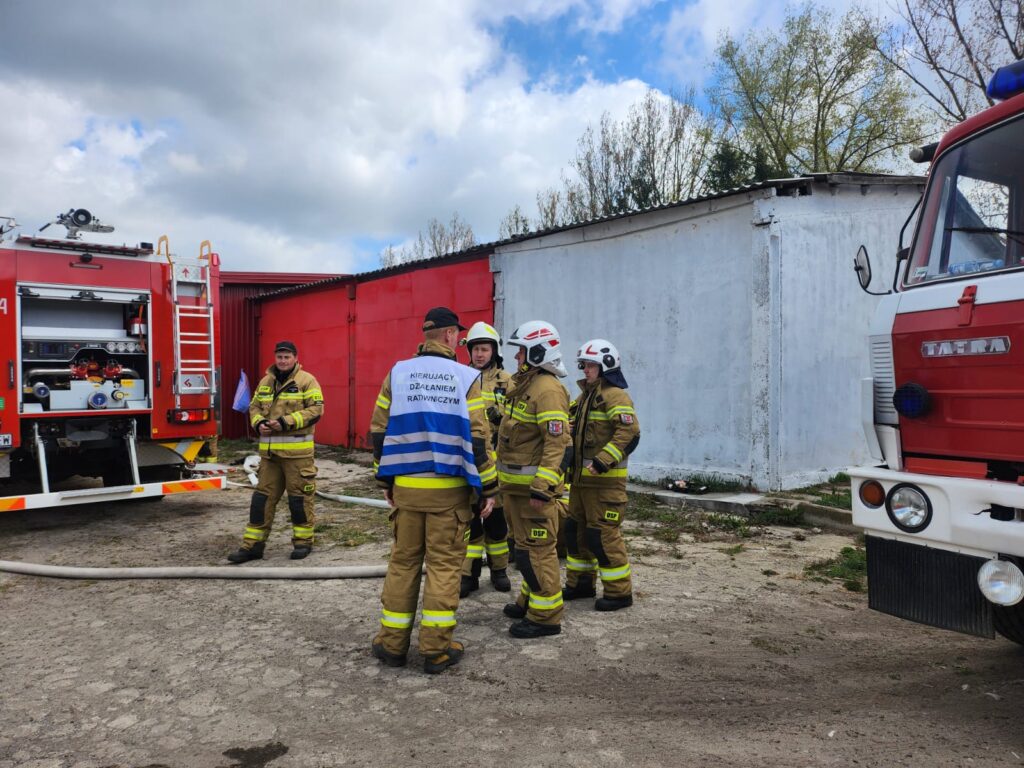 Na zdjęciu są strażacy w strojach ochronnych, z wozem strażackim. Tło to biały budynek i czerwona ściana. Niebo jest pochmurne, ale jasne.