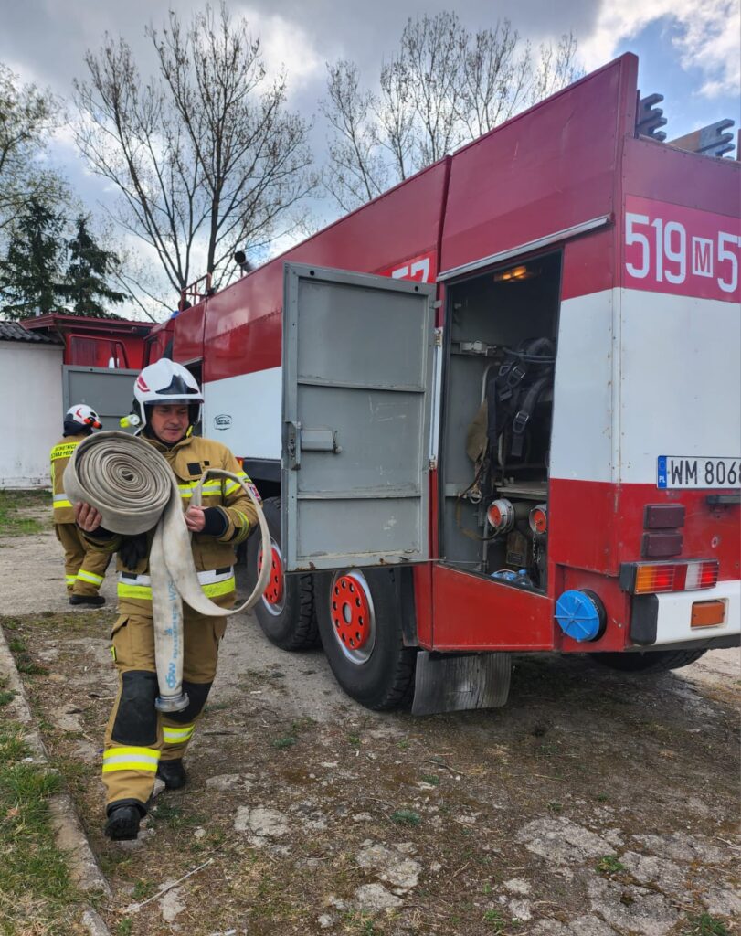 Strażak w żółtym mundurze niesie wąż strażacki. Obok stoi czerwono-biały wóz strażacki z otwartą szafką. W tle drzewa i zachmurzone niebo.
