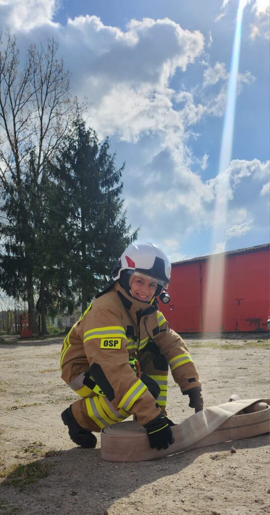 Osoba w stroju strażaka OSP z hełmem na tle niebieskiego nieba i drzew. Uśmiecha się, trzymając zwinięty wąż strażacki. Po prawej stronie widoczny czerwony budynek.