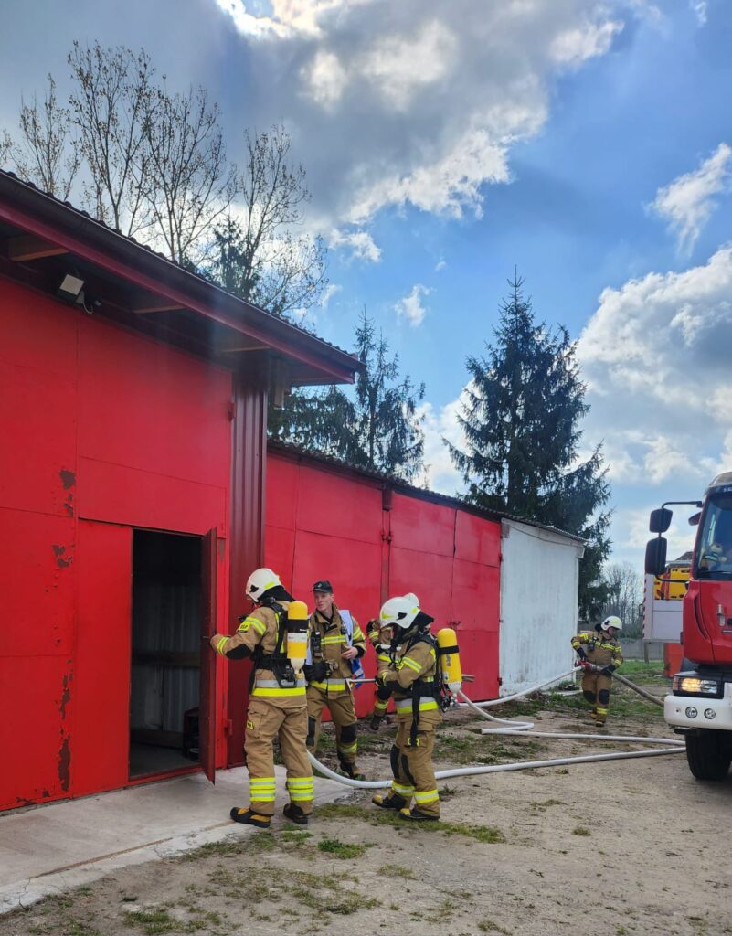 Strażacy w uniformach wchodzą do czerwonego budynku. Na ziemi leżą węże strażackie. Obok stoi wóz strażacki, a w tle widać drzewa i niebo.