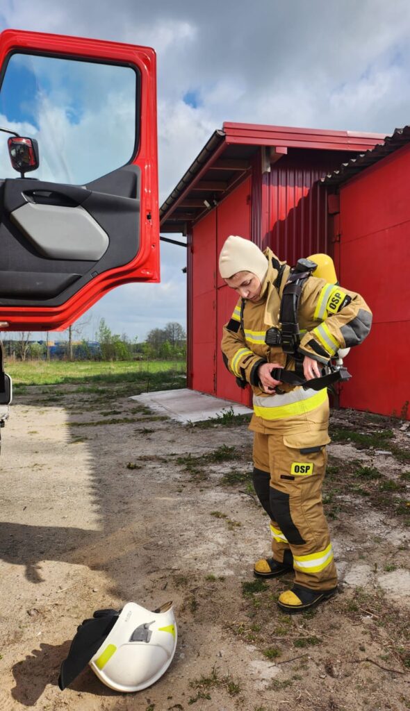 Strażak w beżowym mundurze OSP przy czerwonym wozie strażackim zapina pas. Na ziemi leży biały hełm. Tło to czerwony budynek i niebo.