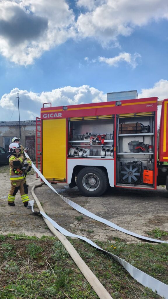 Strażak w stroju ochronnym rozwija wąż strażacki obok czerwono-żółtego wozu strażackiego. Na tle pochmurne niebo oraz zieleń trawy. Wóz strażacki ma otwarte boczne schowki z wyposażeniem.