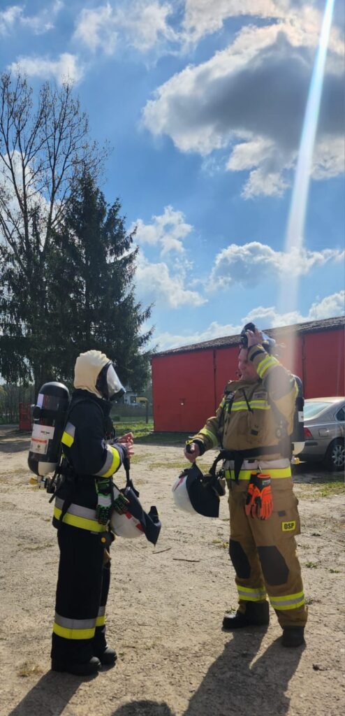 Dwóch strażaków w pełnym umundurowaniu rozmawia na tle nieba z chmurami i czerwonego budynku. Jeden trzyma hełm, a drugi ma maskę na twarzy. W oddali znajduje się samochód i drzewa.