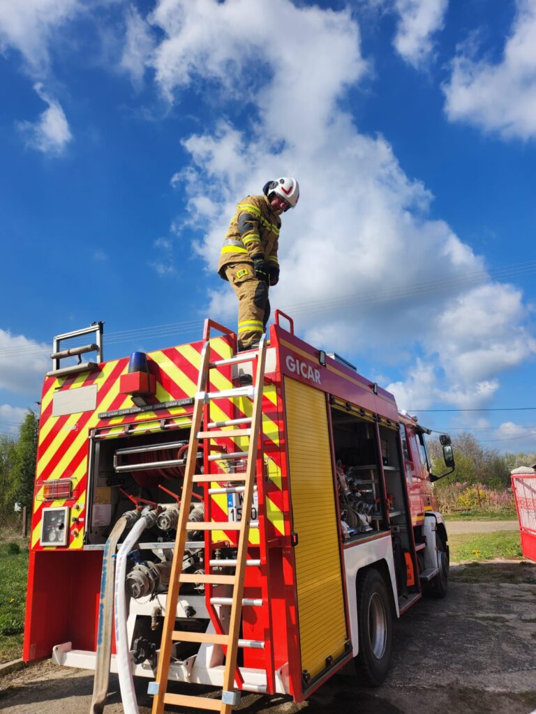 Na zdjęciu strażak na wozie strażackim stoi na drabinie. Pojazd jest czerwony z żółtymi elementami. Tło stanowi błękitne niebo z chmurami.