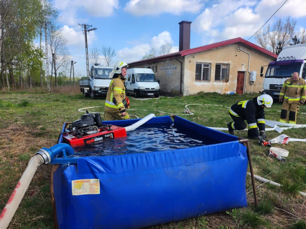 Strażacy w żółtych strojach przy niebieskim zbiorniku wody, na tle wiejskiego domu. Obok białe ciężarówki. Trawa i drzewa wokół.