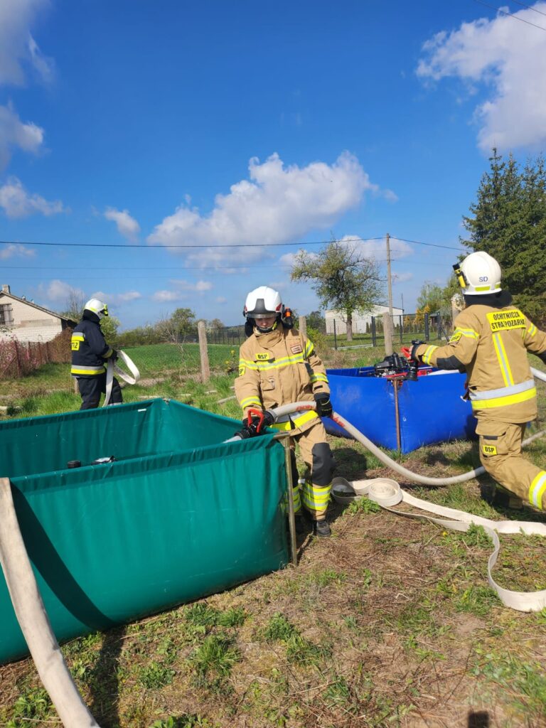 Na tle nieba, trzech strażaków w żółtych uniformach pracuje przy zbiornikach z wodą, jeden zielony, drugi niebieski.
