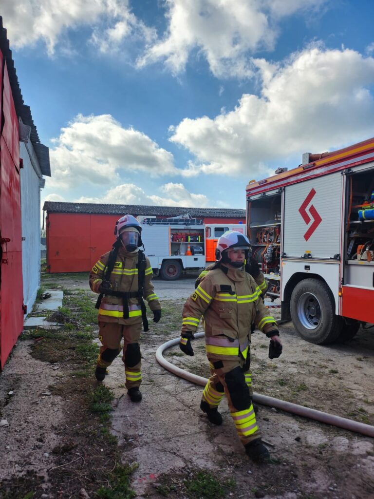 Dwaj strażacy w pełnym umundurowaniu idą obok czerwono-białych wozów strażackich. Na ziemi wąż strażacki, niebo jasne z chmurami. Obok znajduje się czerwony budynek.