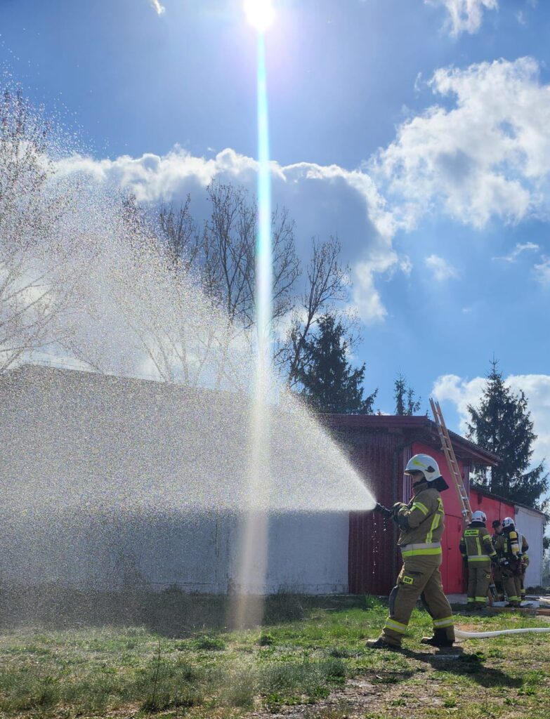 Strażak w mundurze leje wodę na budynek. Słońce oświetla scenę, niebo jest niebieskie z chmurami. W tle czerwona ściana i drabina.